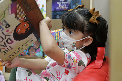 A small child wearing a mask sitting on a beanbag chair holding a book