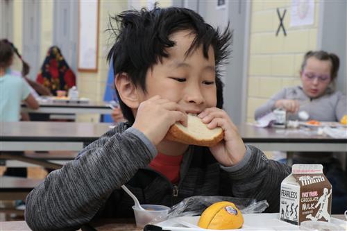 A boy eats a sandwich with fruit and milk