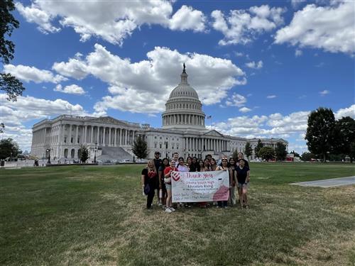 A group of about 15 students and sponsors hold an Educators Rising Banner in front of the U.S. Capitol Building